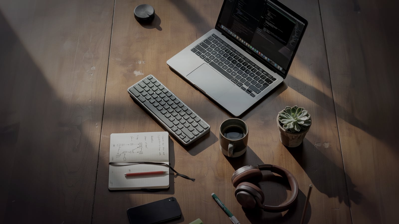 Vista cenital del setup de trabajo SITECH: laptop con código dark theme, teclado mecánico, café, planta, libreta con diagramas